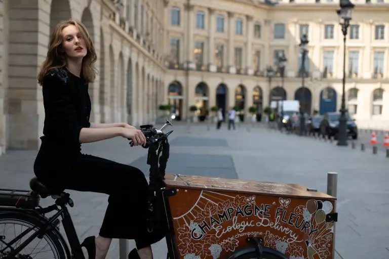 Model in black velvet couture suit with embroidered epaulettes posing beside a champagne delivery cart at Place Vendôme in Paris, by Saskia ter Welle