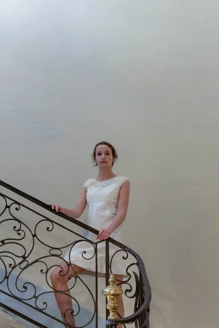 Short white couture dress with all-over pearl embroidery, pearl-accented neckline and feather details, photographed on the staircase of Campredon Art + Image during the opening of the exhibition “Rêves”, by Saskia ter Welle