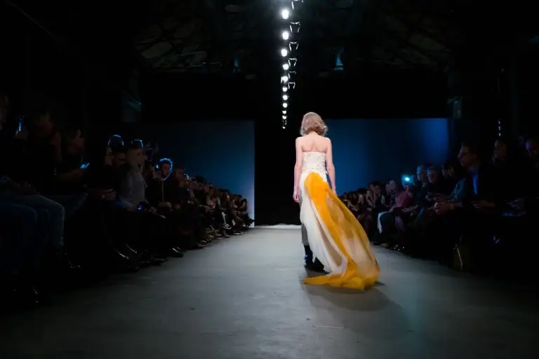 Back view of couture bridal finale outfit with off-white high-waisted trousers, hand-embroidered corset and flowing orange-white train, from Saskia ter Welle’s “Come Fly with Me” show at Amsterdam Fashion Week