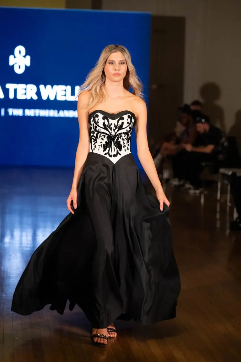 Model walking the catwalk in an off-white and black asymmetrical evening gown with Al Aire embroidery in black velvet and a flowing silk skirt.